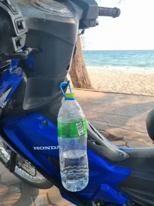 A water bottle that has a handle on it. The handle is hooked onto a hook accessory that is mounted on the dashboard area of a motorbike. The hook accessory gives riders a place to hold things that they purchas, as the lack much storage space on a motorbike.
