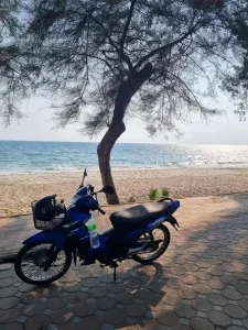 A motorbike (scooter) parked on a sidewalk along a beach in Thailand. The beach is in the background. There are no people in the image. The motorbike is an Honda Wave which is an semi-automatic. This means that the rider switches gears manually without the use of a hand clutch.