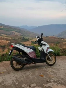 A motorbike (scooter) parked on a back-country road in Thailand, with rolling hills in the background. There are no people in the image. The motorbike is an Honda Click, which is an automatic (not manually or semi-automatic). This means that the rider never switches gears manually - the motorbike has a CVT automatic transmission.