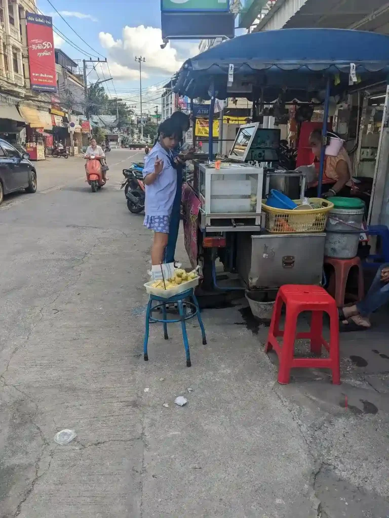 A motorbike-mounted food stall along a road in Thailand. Two people are ordering food. The food cart attached is a metal side cart with cooking setup built onto it. It has a glass container for a food storage area, and has a canopy. This style of food cart is portable, and all the seller needs to do is ride to their selling spot and leave at the end of the day.