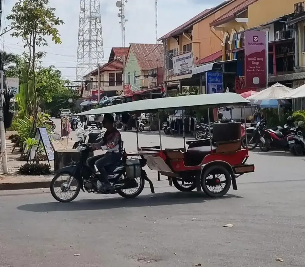 A remorque (French word for wagon) taxi riding down the road in Kampot, Cambodia. It consists of a motorbike that is towing a two-wheeled wagon with seating and a canopy. It has two bench seats - one looking forward, and another one looking backward. Each bench seat fits two people, so four people total can ride in this taxi. This attached wagon-style transportation is unique to Cambodia - it can be used as a family vehicle, a taxi, and sometimes even for moving goods. The motorbike is a Honda Dream in this image.