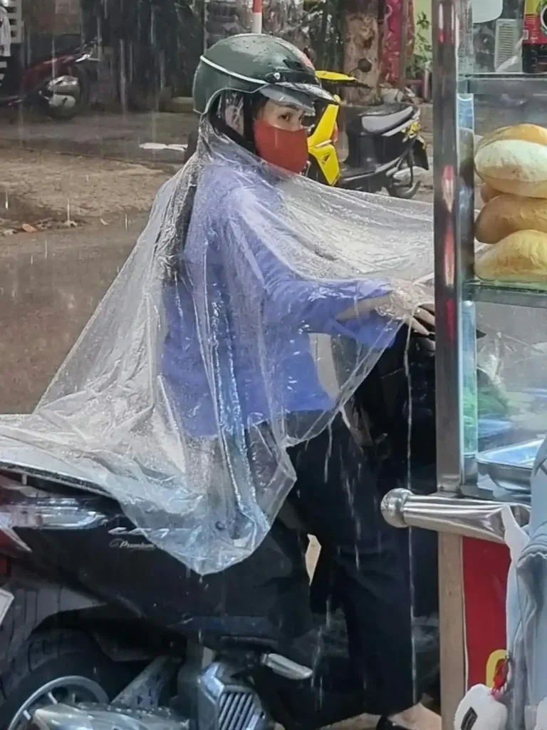 A scooter rider wearing a clear poncho while riding in the rain. The rider is stopped alongside the road in Vietnam purchasing a bahn mi (a Vietnamese sandwich).