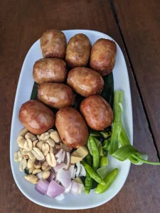 Nine grilled fermented pork Thai sausages. This is called Naem. They are chunky and thumb-sized in length. You can eat them as they are, or with any or all of the condiments included - sliced onion, peanuts, and chilli peppers.