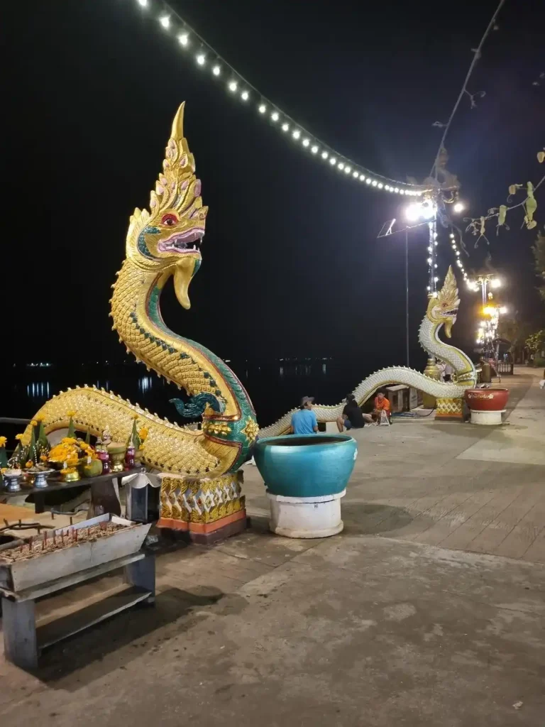 Two naga, a mythical creature that resembles a snake, statues are on each side of a set of stairs that lead down to the Mekong river in Thailand. The statues are intricately decorated and mostly golden colored.