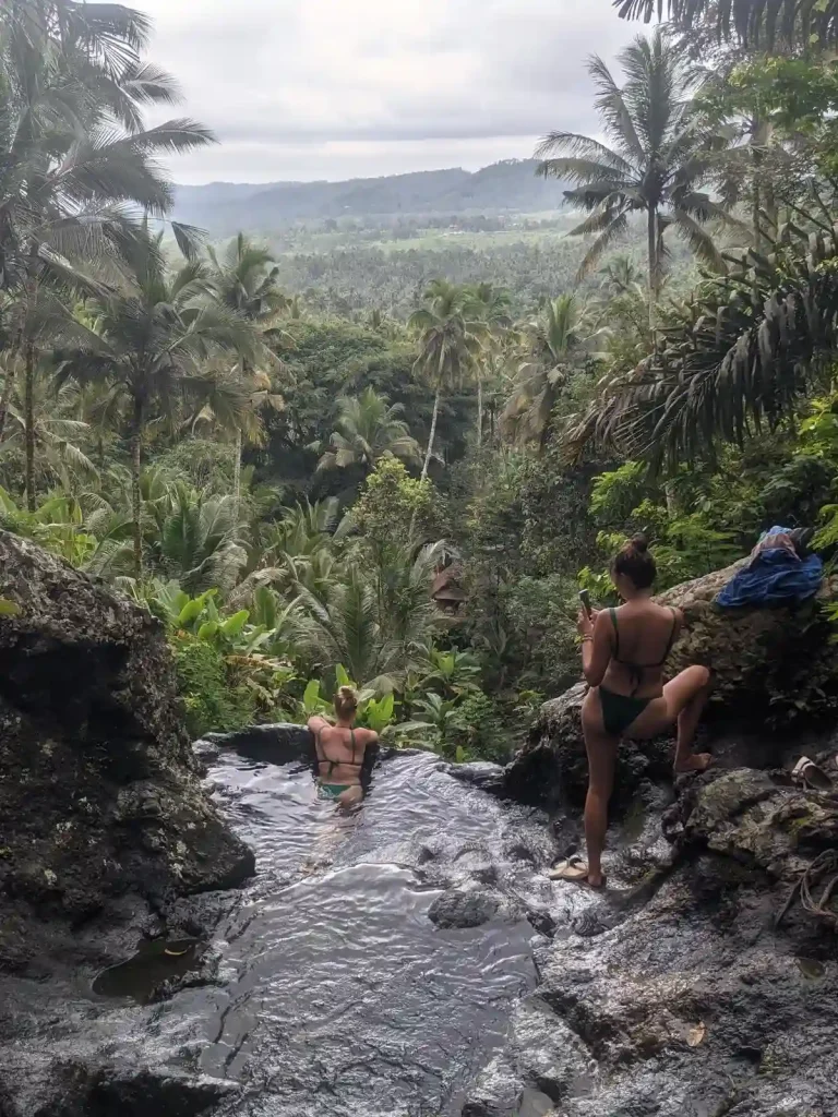 A pool on a waterfall with a view in Bali, Indonesia. At the very peak top of a waterfall with a small natural swimming pool, located at the point where the water falls down. The image shows the water a the bottom half, and the view on the upper half consisting of a forest of palm trees and mountainous region in the background. A female is enjoying the small natural pool that has a rocky protective barrier to prevent them from falling over. The open for the water flows around the barrier. Another female is taking a picture, using their cell phone, of he friend inside the pool.