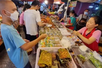 A row overlooking a long table full with various food choices at a bustling food night market in Thailand. A seller is handing back change to a customer who purchased food, holding it in a plastic bag to-go. There are several other people in the background. There is a small walkway (about two people wide) with tables all along each side of the walkway. This image focus on the tables on the right-hand side.
