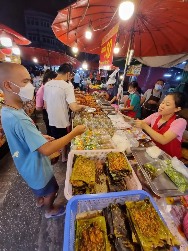 A row overlooking a long table full with various food choices at a bustling food night market in Thailand. A seller is handing back change to a customer who purchased food, holding it in a plastic bag to-go. There are several other people in the background. There is a small walkway (about two people wide) with tables all along each side of the walkway. This image focus on the tables on the right-hand side.