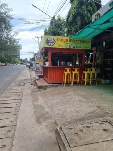 A simple and humble outdoor café along the road in Cambodia. Typically, people get coffee to-go from this style of cafes. It's shape is boxed (about six feet wide, six feet long, and six feet tall) and made of steel. It has a counter/bar that goes around it's perimeter with four chairs. It is in front of a building and takes up the entire sidewalk, blocking it.