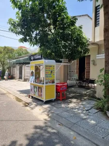 A simple and humble outdoor café cart along the road in Vietnam. It resembles a fancier version of a lemonade stand in the USA. It is located on a sidewalk in front of a house in quiet neighborhood. Typically, people get coffee to-go from this style of cafes. It has pictures of the drinks it serves and a menue with prices at roughly fifty US cents.I t is small, and the shape is boxed (about three feet wide, two feet deep, and six feet tall) and made of steel.