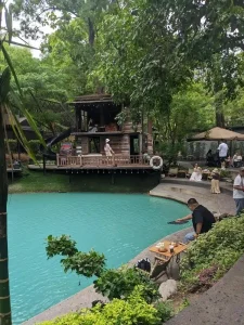 A café with outdoor seating around a small retention pond, filled with greenery - plants and trees. The pond is cleaned and has a ocean-green/blue color. There is a small two-story cabin to sit, too. Many customers are sitting around with drinks of coffee and/or tea.