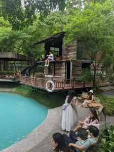 A café with outdoor seating around a small retention pond, filled with greenery - plants and trees. The pond is cleaned and has a ocean-green/blue color. There is a small two-story cabin to sit, too. This image shows the cabin up close. Customers are sitting around with drinks of coffee and/or tea, and taking pictures.