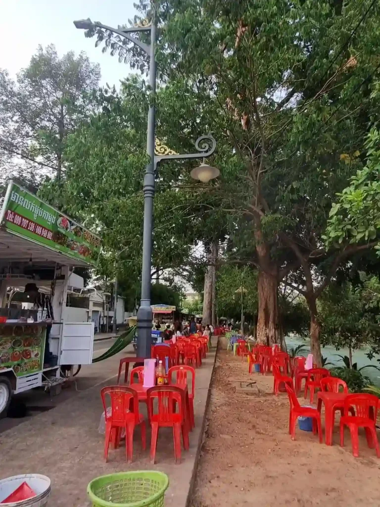 Plastic tables and chairs set up along the river in Siem Reap, Cambodia. This seating is on a small plot of land between the river and sidewalk, with some seating also on the sidewalk. Next to the sidewalk are food trucks and stalls parked along the street, in which customers can order food at, and the seller will cook it within their stall. Keep in mind that walking on sidewalks in Southeast Asia can be challenging as they are often used by local businesses as an extension of their business.