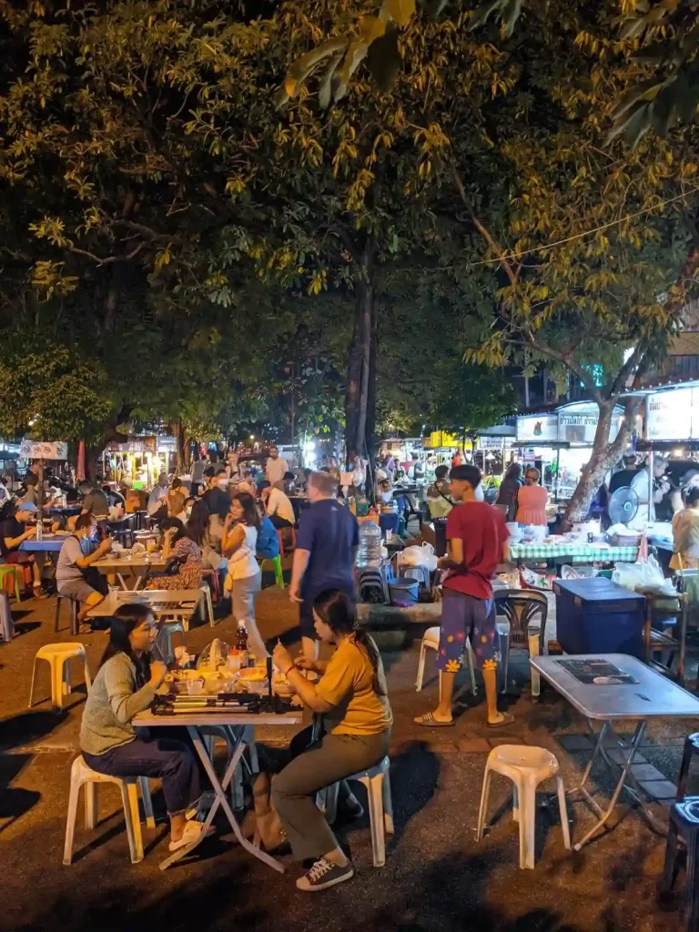 Many tables and chairs setup on a concrete area with many food stall vendors in Chiang Mai, Thailand. This shows a bustling nightlife, and displaying the food culture of Southeast Asia. There are many patrons eating. It is night time in the image, and the lighting is not bright, which gives it a relaxing vibe.