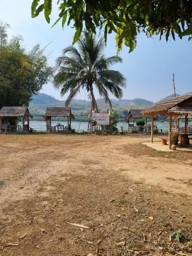 An outdoor restaurant in Thailand with several small huts, each with a table inside and seating. This is located along the Mekong river in rural Thailand. A couple of palm trees are on the property.