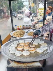 A woman with an enclosed food cart is pan frying chive cakes on the side of a narrow street in Cambodia. The cart has several sauces to choose from. The pan is large - about twenty inches in diameter - and is heated underneath with wood fire stove burning small pieces of chopped wood. In the background, a customer is purchasing some food.