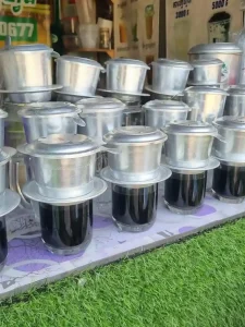 A Phin coffee brewing filter (pronounced "feen" as in the ending of the wording "caffeine") resting on top of a glass cup, brewing coffee. This image shows several cups being brewed in batch in preparation for future customers at a roadside cafe. This coffee filter is filled with ground coffee beans, and hot water is added inside. It uses gravity to slowly percolate (drip) hot water through the coffee grounds. At the bottom of this filter, the hot coffee drips out into a cup. This brewing method was introduced to Vietnam and Cambodia by the French during the colonial occupation time period.