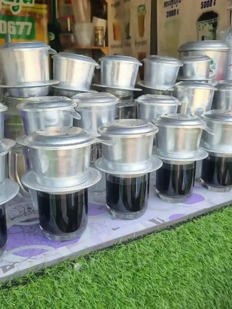 A Phin coffee brewing filter (pronounced "feen" as in the ending of the wording "caffeine") resting on top of a glass cup, brewing coffee. This image shows several cups being brewed in batch in preparation for future customers at a roadside cafe. This coffee filter is filled with ground coffee beans, and hot water is added inside. It uses gravity to slowly percolate (drip) hot water through the coffee grounds. At the bottom of this filter, the hot coffee drips out into a cup. This brewing method was introduced to Vietnam and Cambodia by the French during the colonial occupation time period.