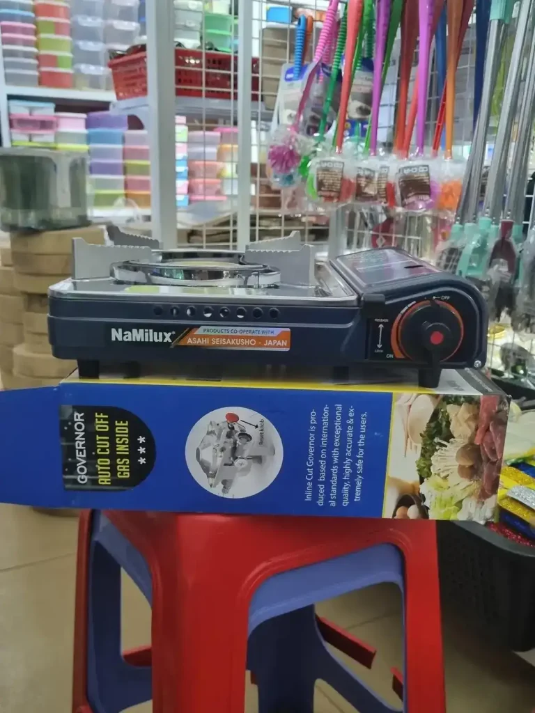 A side view of a portable gas cooker for sale in a store in Southeast Asia. This image shows how tall the cooker and it's stove section is. The gas canister is inserted on the side, and it's compartment takes up the length of the cooker. It has a stove with holders in all four corners to set pans ontop of. The holders are long, meaning they can hold different sizes (small or large) of pots or pans. It is about one and a half feet wide, two feet long, and five inches tall (61 cm by 61 cm by 2.5 cm).