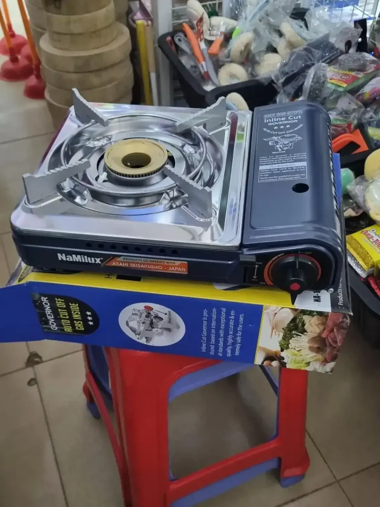A portable gas cooker for sale in a store in Southeast Asia. The gas canister is inserted on the side, and it's compartment takes up the length of the cooker. It has a stove with holders in all four corners to set pans ontop of. The holders are long, meaning they can hold different sizes (small or large) of pots or pans. It is about one and a half feet wide, two feet long, and five inches tall (61 cm by 61 cm by 2.5 cm).