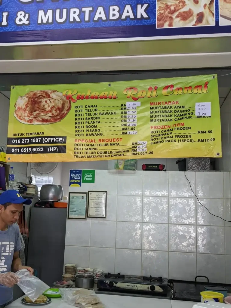 A price list for food at a small restaurant in Malaysia. This restaurant serves Roti Canai, which is a southern Indian-style dish (Malaysia has a large population Tamil-ethnicity Indians from Southern India). The menu shows a roti, which is a soft, round, flat bread that has a unique flavor, which is accompanied by a chosen type of sauce. Meat can be added. Canai is pronounced the same as "Chennai", the large southern Indian city.