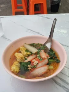 A bowl of egg noodles with red grilled pork, spinach, and a few dumplings inside. An asian-style soup spoon is sticking out of the bowl. The scenery is outdoors, and a couple of small plastic stool seats can partially be seen in the image.