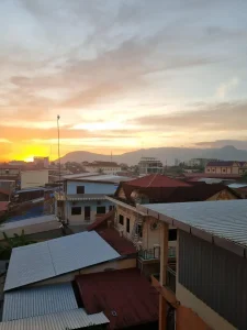 A rooftop view of a sunset. The sun is setting behind some knobs (in between a hill and a mountain). Nearby, there are buildings a story or two below, in which their corrugated steel roofs can be seen.