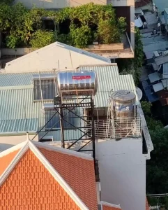 A rooftop view looking down at the rooftops below in Vietnam. The buildings, a few stores below, have roofs made of clay tiles and corrugated steel sheets. A cistern (large water storage) is mounted on their rooftops, and is plummed to their home. This is very common in Southeast Asia - to have a rooftop water cistern with gravity pressure fed throughout the home. It is heated by the sun. Some homes have electrical on-demand water heaters within each bathroom shower for heated water.