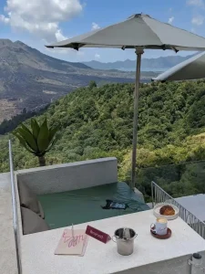 Seating at a rooftop café overlooking Mount Batur in Bali, Indonesia. There is only one seat in this image, which is large seat fitting up to two people. It is cushioned, and has an umbrella for shade. Attached to the seat, is a large table on the right-hand side with a café late and croissant. Mount Batur is on the left-hand side of the seat.