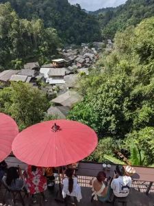 Seating at a rooftop café overlooking a traditional village in a mountainous area of northern Thailand. There is a long bar at the edge of the rooftop with seats that face the village, below. Umbrellas are near the seating for shade. Many customers are enjoying drinks such as coffee and milk teas.