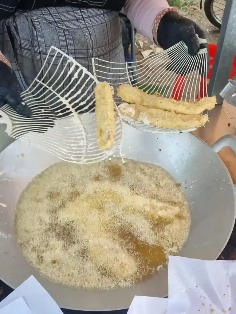 A food seller's kiosk along a road in Cambodia, in which they are preparing deep fried bananas. They are fishing-out the bananas out of a pan of oil using a tool. This is a common snack throughout Southeast Asia. Bananas are peeled, sliced, covered in a battered, and then deep fried.