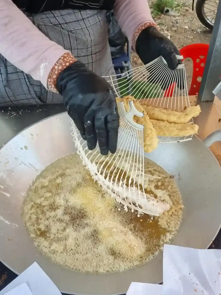 A food seller's kiosk along a road in Cambodia, in which they are preparing deep fried bananas. They are fishing-out the bananas out of a pan of oil using a tool. This is a common snack throughout Southeast Asia. Bananas are peeled, sliced, covered in a battered, and then deep fried.