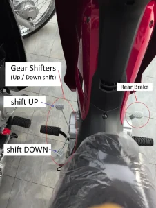 An image of a semi-automatic motorbike. There is text with arrows pointing at the controls of a semi-automatic motorbike. This image highlights the locations of the gear shifters and rear brake. The gear shifters (one for shifting up, and another for shifting down) are on the left-hand side foot peg, and the rear brake is on the right-hand side foot peg.