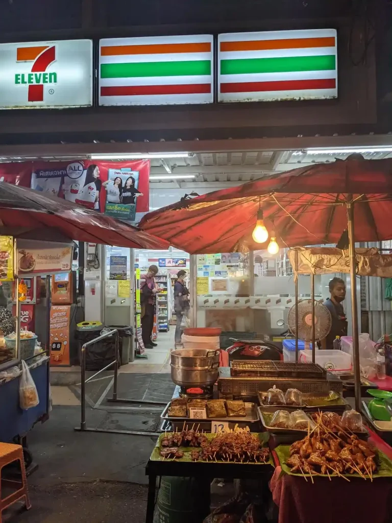 A Seven Eleven convenience store in Thailand with food stalls outside the entrance. Food includes a few different grilled meats and a few Thai dishes packaged in plastic bags.
