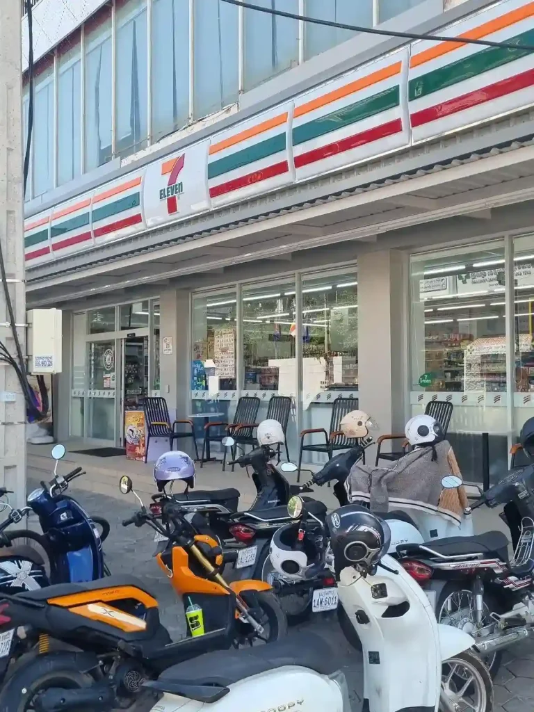 A Seven Eleven convenience store in Southeast Asia with motorbike (scooter) parking on the sidewalk outside the entrance. There is seating and tables on the patio outside the store for relaxing.