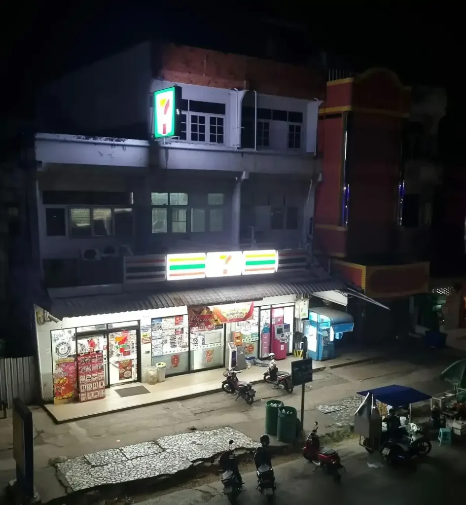 A Seven Eleven convenience store in Thailand with motorbike (scooter) parking on the sidewalk outside the entrance. This image captures the entire three-story building, which has the Seven Eleven on the ground floor, three small apartments on the second floor, and two small apartments on the third floor. A street food vendor is parked outside on the street selling food.