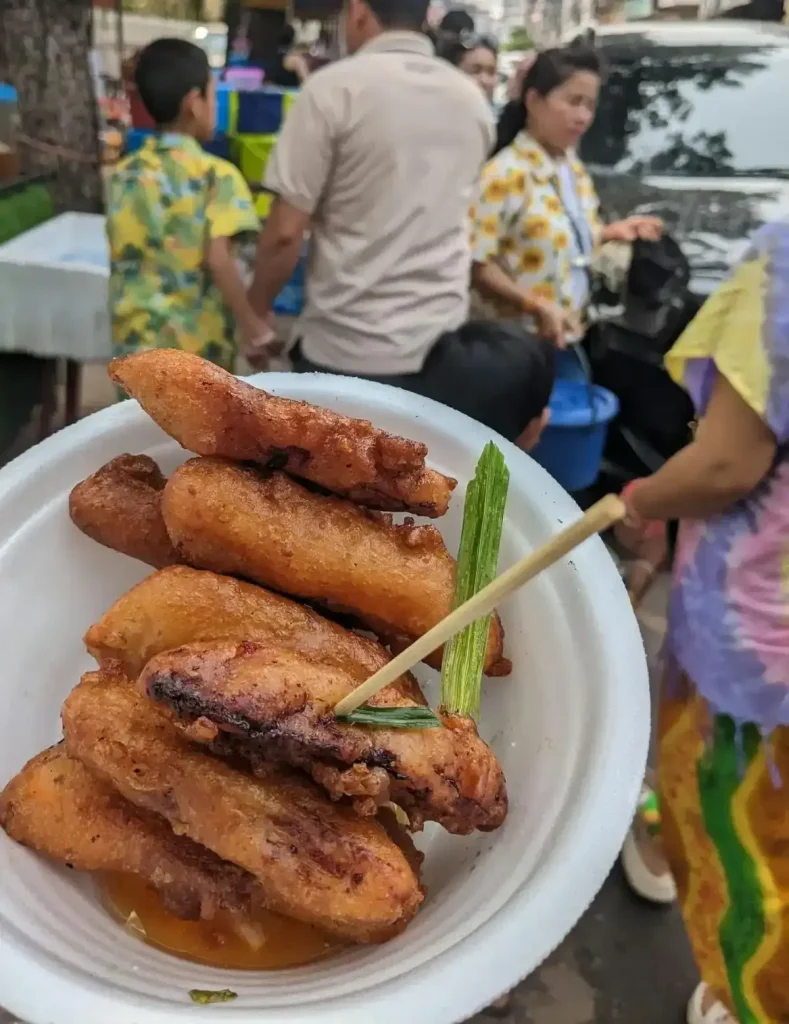 Deep fried bananas, the short and chunky variant. This a common snack throughout Southeast Asia. A wooden stick, like a large tooth pic, is stuck into one of the banana slices and used as a fork. Bananas are peeled, sliced, covered in a battered, and then deep fried. Deep fried bananas come in a few different variations. The ones in this image are the short, chunky, version. This image was taken in Laos during Songkran holiday, so in the background are local Laos people wearing festive Songkran shirts, in which are very similar to a Hawaiian shirt. This shirt is not a traditional local shirt, but has been made popular with the evermore commercialization of the Songkran holiday in Cambodia, Laos, Thailand, and globalized cities such as New York City.