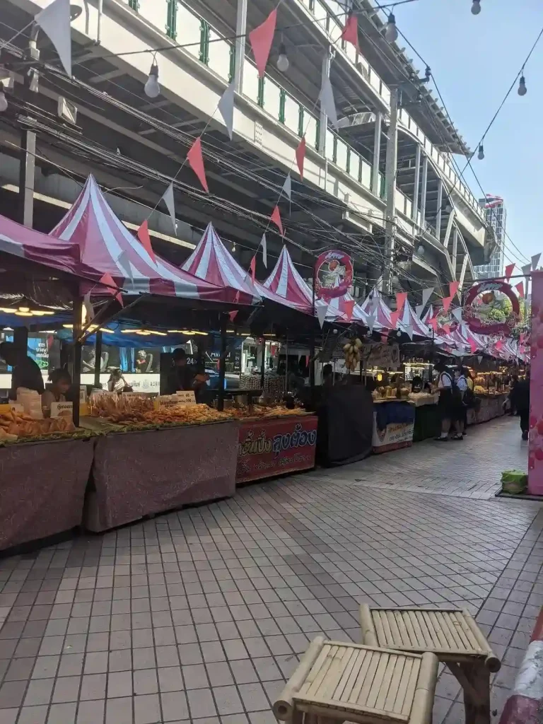 A line of food stalls with canopies, side-by-side, down a sidewalk. This is located below the tracks of an elevated subway line (aka metro or tube) in Bangkok, Thailand. There is plenty of walking space, and the presentation is orderly.