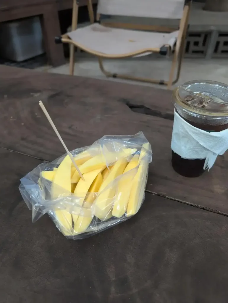 A bag of sliced not-fully-ripe mango on a table next to an iced Americano coffee at a café in Southeast Asia. There is a stick, like a large tooth pic, that is stuck into a piece of the mango. It serves as a fork. Not-fully-ripe mango is crunchy, much like an apple. It is a popular way to eat mango in Southeast Asia. A benefit is that the sellers have more time to sell it before it ripens, then goes bad.