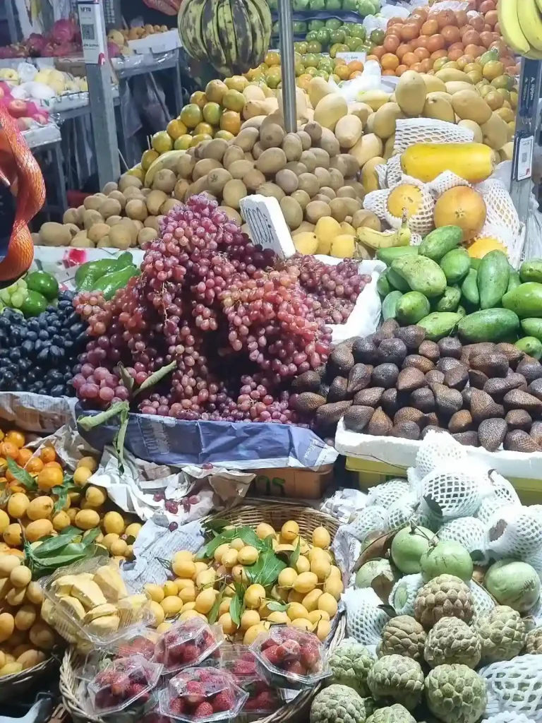 A close up image of various fruit being sold at a stall in Southeast Asia. Jack fruit, mangos, passion fruit, oranges, purple and dark purple grapes, bananas, and more are sold here.