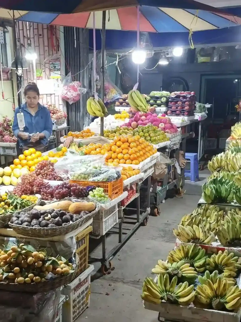 A stall in Southeast Asia selling a variety of fruit. Bananas, passion fruit, dragon fruit, oranges, green and purple grapes, and apples are sold here. The stall is partially outdoors with an umbrella over the fruit. A seller is standing around waiting for customers.