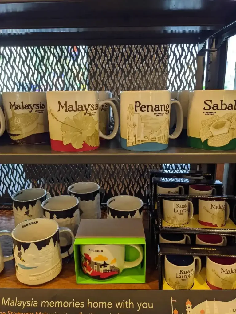 A rack of coffee mugs for sale at Starbucks coffee shop in Malaysia. Some mugs are stylized with the name of the country, Malaysia, on it. Other mugs have the name of a Malaysian city. Cities include: Penang, Sabah, Kuala Lumpur, Sarawak, and Kuching.
