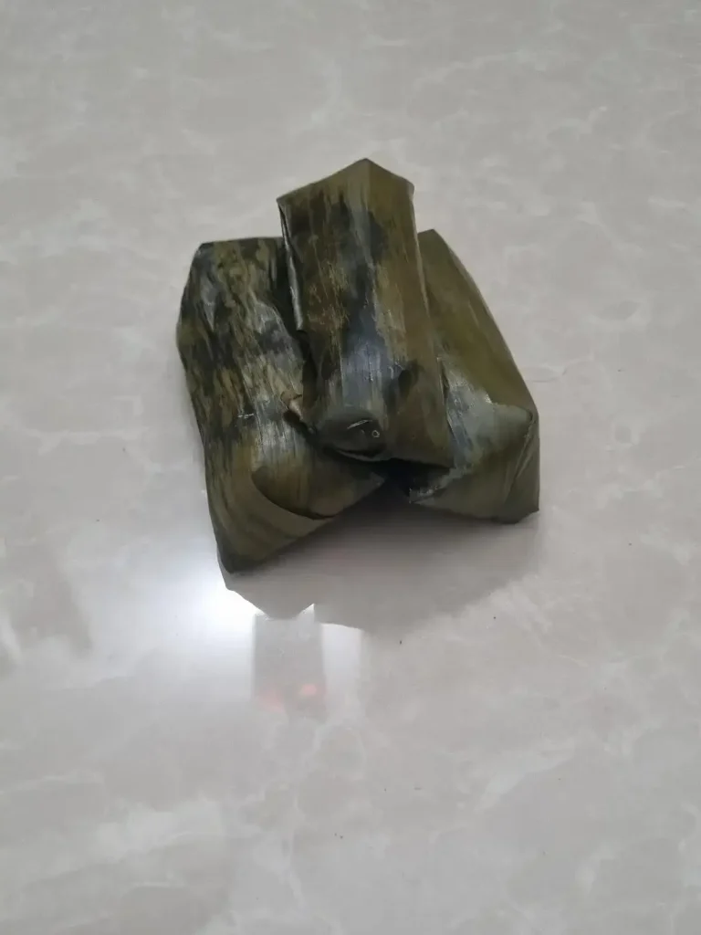 Three steamed gluttonous sticky rice covered banana snacks, each wrapped in banana leaf.