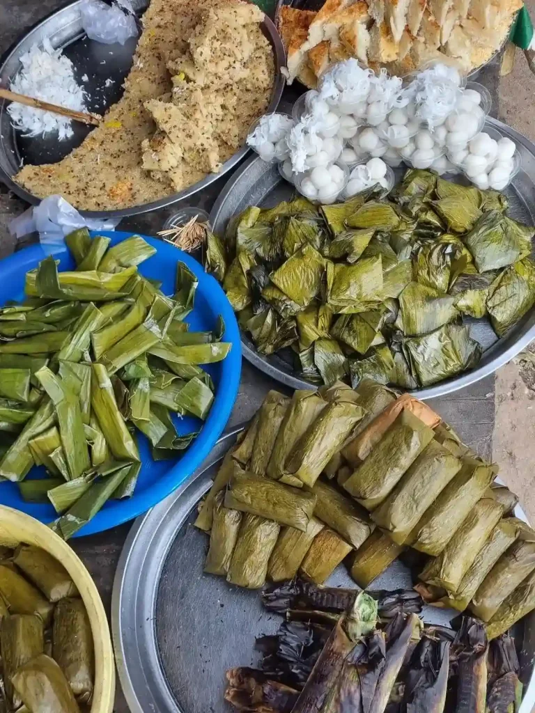 A variety of steamed and/or grilled gluttonous sticky rice covered banana snacks, each wrapped in banana leaf, for sale at a market in Southeast Asia. There are several variants of different shapes and banana types.