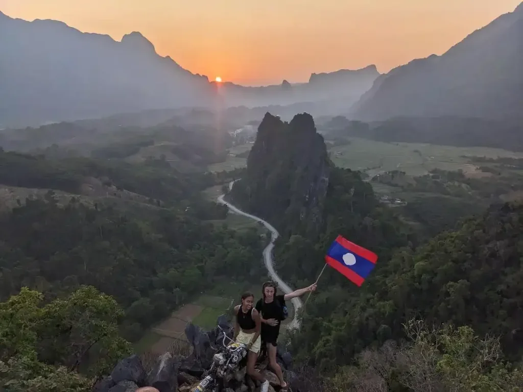 A popular and well-known sunset viewpoint in Vang Vien, Laos. It requires hiking up a mountain. At the top is a view of the land in three-hundred and sixty degree direction of flat land surrounded by mountains. This viewpoint has a motorcycle with a Laos flag for posing in pictures with. The sunset is visible at the top edge of one of the mountains, before it gets covered by it.