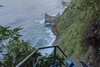 Steep metal stairs mounted on a very high cliff that leads from the top to the ocean. This image is mid-way on the stairs, showing the near vertical cliff and how it meets the ocean water at the bottom.