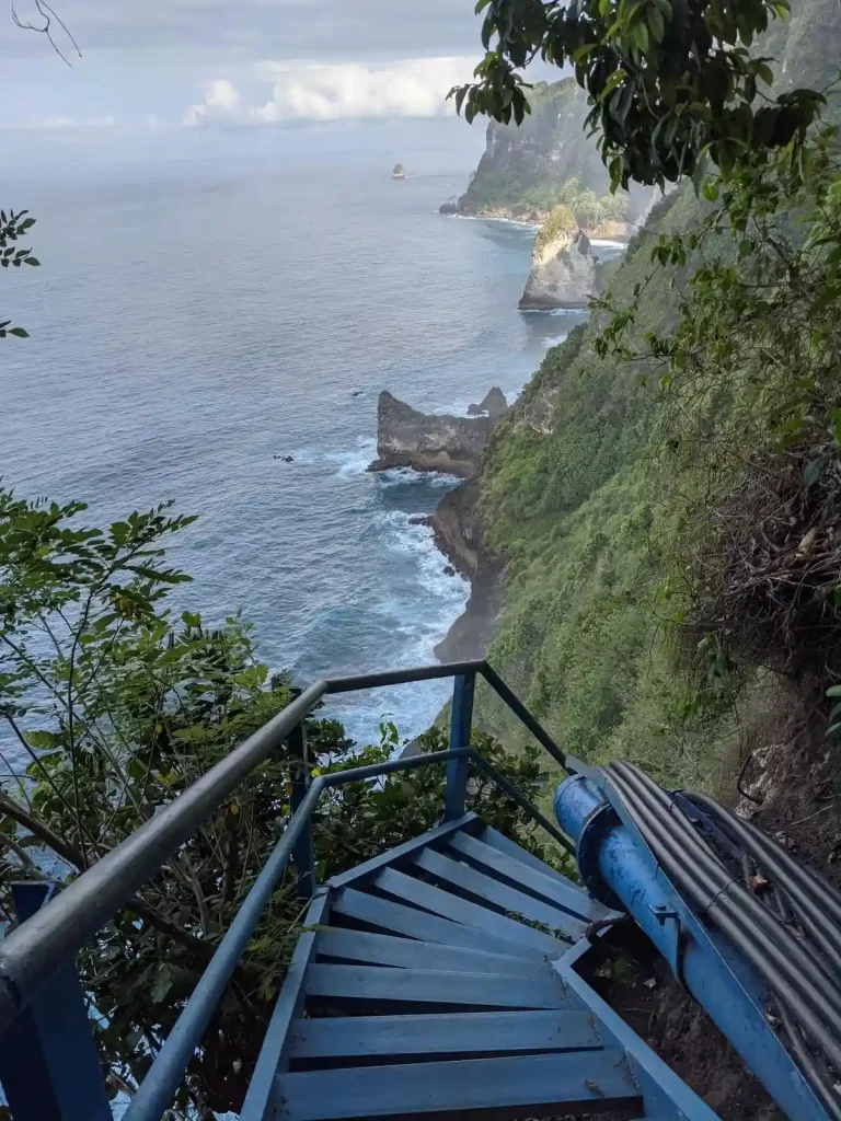 Steep metal stairs mounted on a very high cliff that leads from the top to the ocean. This image is mid-way on the stairs, showing the near vertical cliff and how it meets the ocean water at the bottom.