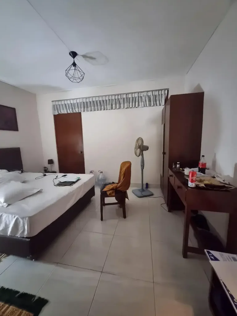 A furnished studio room rented monthly in Southeast Asia. The image shows a bed with sheets, the front door, a wardrobe along one wall, a chair and desk along the wall. The floor is tile, which is very common in Southeast Asia.