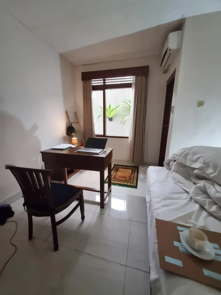 A furnished studio room rented monthly in Southeast Asia. The image shows how the tenant has a table with laptop on it and chair faced looking outside a window. The split-duct airconditioning unit is above the bathroom door. There is a muslim prayer mat, which is fitting due to the location being Indonesia. The image also shows the edge of the bed with sheets. The floor is tile, which is very common in Southeast Asia.