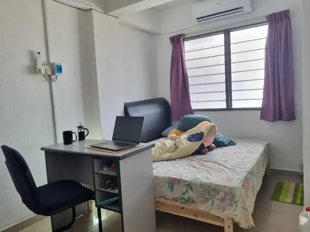 A furnished studio room rented monthly in Southeast Asia. The image shows how the tenant has a table with laptop on it and chair faced looking outside a window. The table is at the end of a queen size bed. On the other side of a bed is a window with opaque film on it making it impossible to see outside. The split-duct airconditioning unit is above the window. The image also shows an electrical meter, which is used to measure each the tenants electricity usage and calculate monthly electric fee based on it.