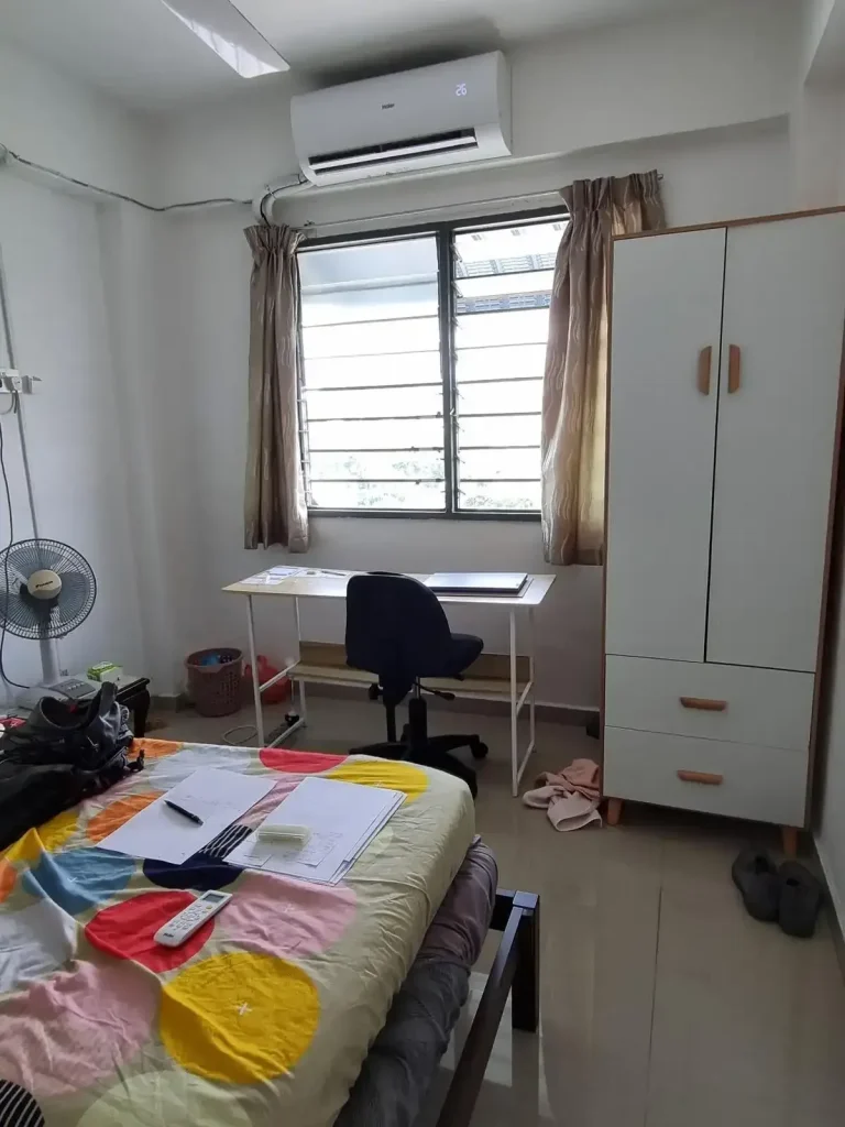 A furnished studio room rented monthly in Southeast Asia. The image shows how the tenant has a table with laptop on it and chair faced looking outside a window. The table is along the wall in front of the window. to the side of the desk is a small and wardrobe storage. To the other side is an airfan. The split-duct airconditioning unit is above the window. The image also shows the edge fo a twin-sized bed.